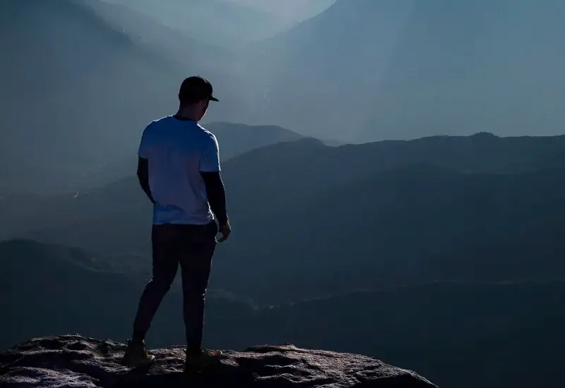 A man stands on a rocky ledge looking out over layers of misty mountains at first light